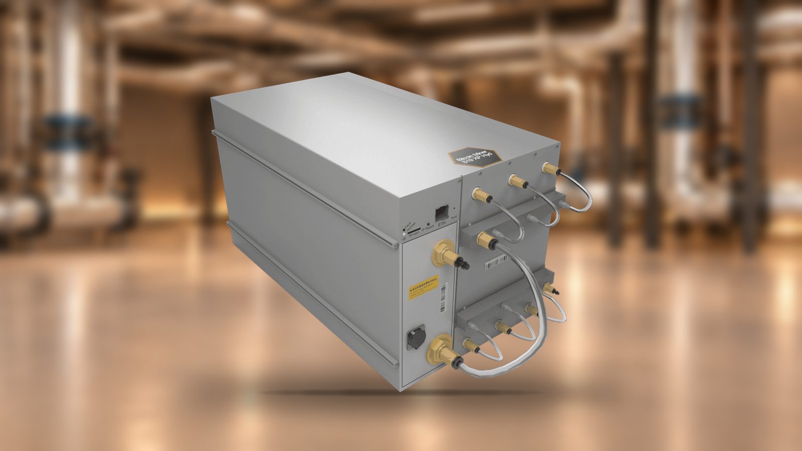 An Antminer S21e XP Hyd with its brass coolant fittings, in front of a softly blurred plant room with silver insulated pipework