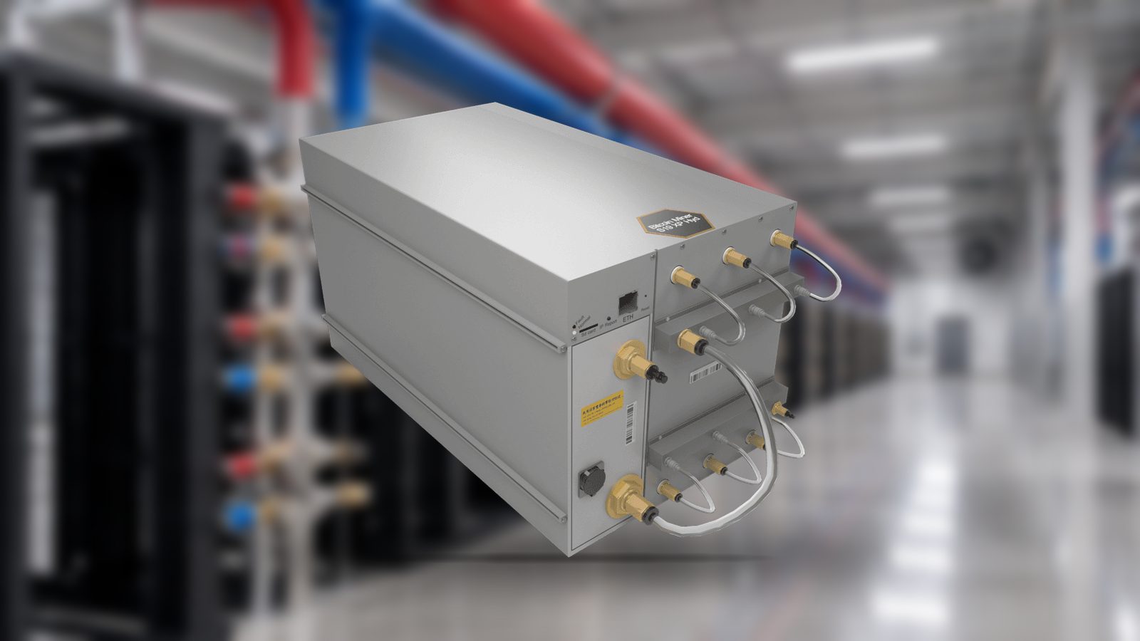 A Bitmain Antminer S19e XP Hyd photographed in the corridor of a hydro-cooled data hall, overhead coolant lines feeding a row of manifolds