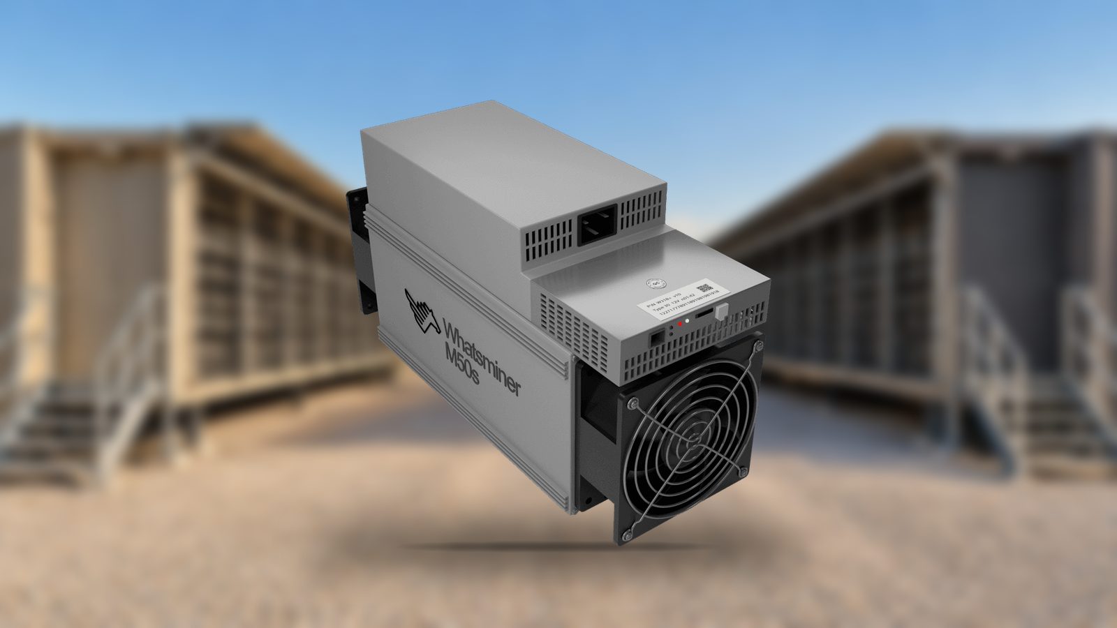 A Whatsminer M50 series air-cooled miner, shown whole, in front of a blurred row of air-cooled mining containers on a gravel lane