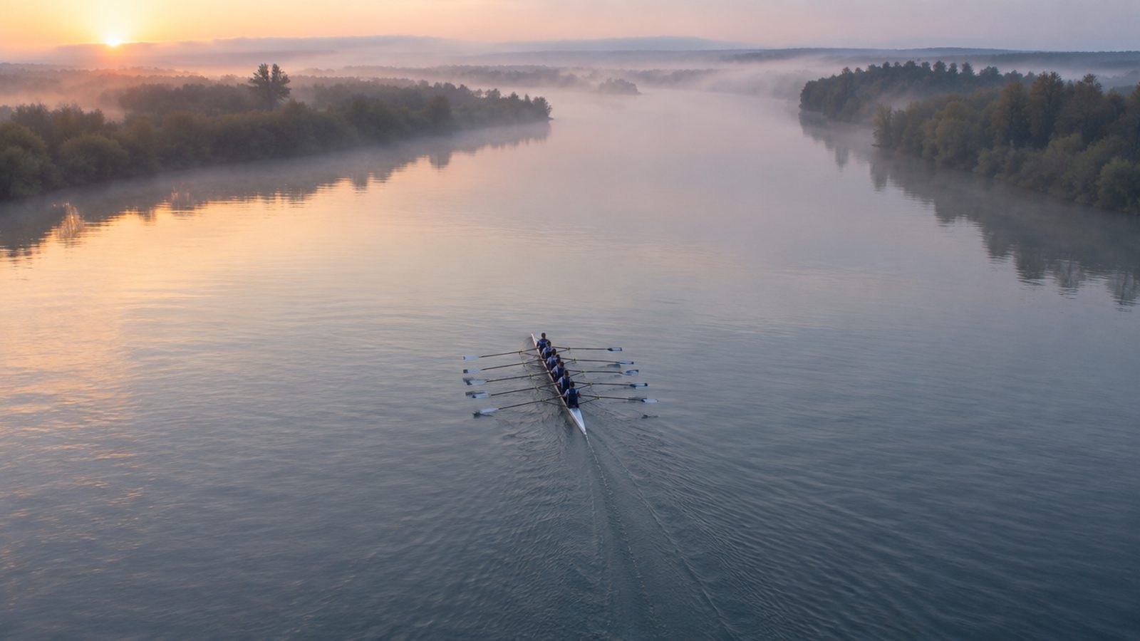 A rowing eight moving down a wide, misty river at dawn, seen from a bridge high above